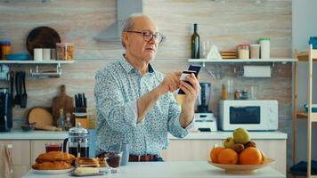 Senior Mann tun online Transaktion mit Telefon App zum Zahlung während Frühstück im Küche. im Ruhestand Alten Person mit Internet Zahlung Zuhause Bank Kauf mit modern Technologie foto
