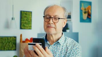 Senior Mann Einkaufen online mit Smartphone und halten Anerkennung Karte während Frühstück im Küche. im Ruhestand Alten Person mit Internet Zahlung Zuhause Bank Kauf mit modern Technologie foto