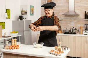 glücklich Senior Mann knacken Eier Über Weizen Mehl während vorbereiten traditionell Rezept. Alten Gebäck Koch knacken Ei auf Glas Schüssel zum Kuchen Rezept im Küche, Mischen durch Hand, kneten foto