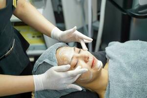 Nahansicht Kosmetikerin bewirbt sich glatt Gesichts- Weiß Maske Sahne und Massage auf Schönheit Frau Herstellung Gesicht Spa im Schönheit Klinik. foto