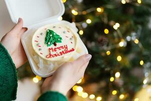 ein Mädchen hält im ihr Hände ein hausgemacht Weihnachten Kuchen mit das Inschrift fröhlich Weihnachten, gegen das Hintergrund von ein dekoriert Weihnachten Baum. foto