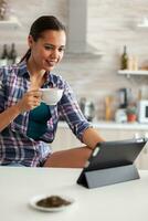 genießen Tasse von Grün Tee während mit Tablette pc im Küche. Arbeiten von Zuhause mit Gerät mit Internet Technologie, tippen, auf Gadget während Frühstück. foto