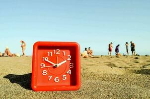 ein rot Uhr auf das Strand mit Menschen im das Hintergrund foto