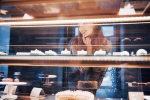 lächelnde Frau in die Kamera durch die Vitrine mit Süßigkeiten und Kuchen im modernen Café-Interieur foto