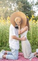 junges glückliches Paar, das sich auf einer Picknickdecke küsst und ihre Gesichter mit einem Sommerhut bedeckt