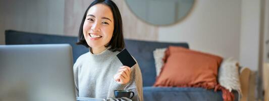 glücklich lächelnd Mädchen mit Anerkennung Karte, bezahlen ihr Rechnungen online auf Computer, tun Einkaufen auf ihr Laptop, Sitzung beim Zuhause foto