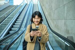 Porträt von schön asiatisch Mädchen, Schüler geht Nieder Rolltreppe im Stadt, sieht aus beim Handy, Mobiltelefon Telefon, Verwendet Telefon, Karte Anwendung, pendelt irgendwo im Stadt, Dorf foto