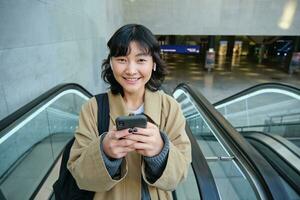 Porträt von süß lächelnd Koreanisch Mädchen, geht oben Rolltreppe, hört zu Musik- im kabellos Kopfhörer und Verwendet Handy, Mobiltelefon Telefon, hält Smartphone foto