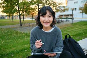 jung asiatisch Mädchen mit Grafik Bleistift und Tablette, sitzt im Park auf Bank, zeichnet Kratzer, tut ihr Hausaufgaben draußen foto