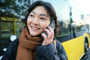 zellular Technologie und Menschen Konzept. stilvoll asiatisch Mädchen Gespräche auf Handy, Mobiltelefon Telefon, macht ein Telefon Forderung, steht in der Nähe von Bus halt und hat Konversation foto