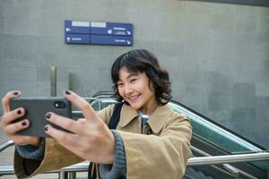 Porträt von stilvoll Koreanisch Mädchen, Schüler nimmt Selfie auf Straße, hält Smartphone und posiert auf es ist Kamera, macht Video auf Tourist Reise, lächelt mit Freude foto