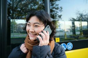 zellular Technologie und Menschen Konzept. stilvoll asiatisch Mädchen Gespräche auf Handy, Mobiltelefon Telefon, macht ein Telefon Forderung, steht in der Nähe von Bus halt und hat Konversation foto