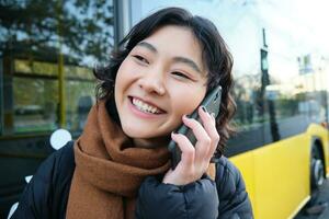 schließen oben Porträt von glücklich asiatisch Mädchen Gespräche auf Handy, Mobiltelefon Telefon, lächelt während spricht zu jemand auf Smartphone, steht in der Nähe von Bus auf halt foto