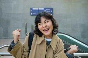 begeistert asiatisch Mädchen steht auf Straße, macht Hurra Feier Geste, sagt Ja, triumphierend, gewinnen und genießen diese Gefühl, Stehen allein draußen foto