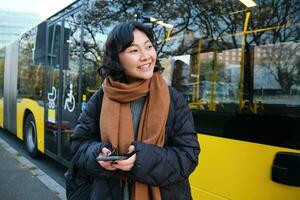 Porträt von Koreanisch Mädchen Kauf Fahrkarte zum Öffentlichkeit Transport online, mit Handy, Mobiltelefon Anwendung auf Bus stoppen, tragen Winter Kleider foto