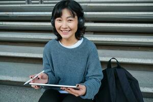 jung asiatisch Mädchen, Schüler im Kopfhörer, funktioniert auf Fernbedienung, Digital Künstler Zeichnung auf Tablette mit Grafik Stift, Hören Musik- im Kopfhörer und Sitzung auf Straße Treppe foto