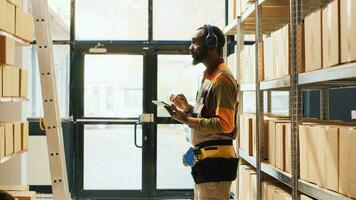 glücklich jung Mann Hören zu Musik- Überprüfung Kisten im Warenhaus Raum, genießen Lieder auf Headset beim arbeiten. männlich Arbeiter Singen und Arbeiten auf Logistik und Inventar mit Tablette. Stativ Schuss. foto