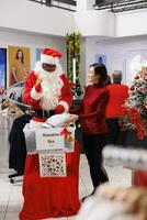 Frau assistieren mit spenden zu Nächstenliebe im Kleidung speichern, Arbeiten mit Santa claus Mitarbeiter zu Verbreitung Freundlichkeit während Dezember Jahreszeit. Klient und Arbeiter Füllung im Spende Box zum Kinder. foto