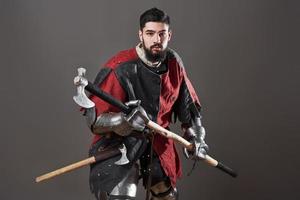 mittelalterlicher Ritter auf grauem Hintergrund. Porträt eines brutalen Kriegers mit schmutzigem Gesicht mit roter und schwarzer Kleidung mit Kettenpanzerrüstung und Streitaxt foto