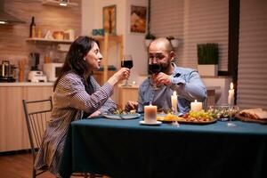 Paar Toasten mit Wein Brille während romantisch Abendessen. entspannen glücklich Menschen klirren, Sitzung beim Tabelle im Küche, genießen das Mahlzeit, feiern Jahrestag im das Essen Zimmer. foto