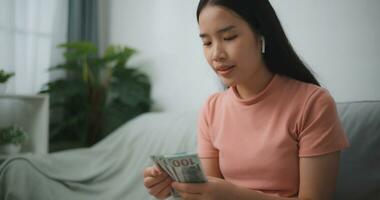 Porträt von glücklich jung asiatisch Frau zählt Kasse Dollar Rechnungen während Sitzung im Leben Zimmer beim Zuhause, Zählen Stapel Kasse Geld Dollar foto