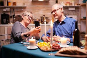 alt Paar mit Telefone im das Küche während romantisch Abendessen. Sitzung beim das Tabelle im das Essen Zimmer , durchsuchen, suchen, mit Telefon, Internet, feiern ihr Jahrestag im das Essen Zimmer. foto