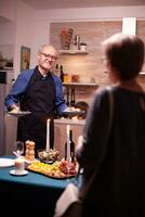 Senior Mann Portion Ehefrau während feiern ihr Beziehung mit lecker und und Wein. Alten alt Paar reden, Sitzung beim das Tabelle im Küche, genießen das Mahlzeit, feiern ihr Jahrestag . foto