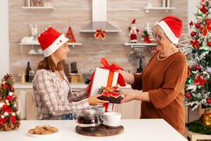 Oma überraschend Enkelin mit Weihnachten Verpackung Geschenk feiern Weihnachten Urlaub zusammen im dekoriert Küche. Kind genießen Winter Jahreszeit Ändern Geschenk mit Band auf es mit Gradma foto