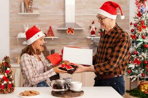 Großvater überraschend Enkelin mit Weihnachten Verpackung Geschenk genießen Weihnachten Urlaub zusammen im dekoriert Küche. Kind feiern Winter Jahreszeit Ändern Geschenk mit Band auf es mit Opa foto