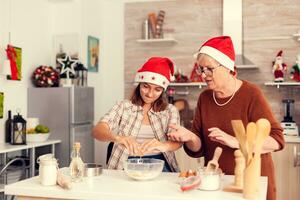 wenig Nichte während Weihnachten Tag Kochen und haben Spaß mit Oma. glücklich heiter freudig Teenager Mädchen Portion Senior Frau vorbereiten Süss Kekse zu feiern Ferien tragen Santa Hut. foto