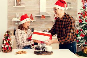 erstaunt heiter Nichte mit Weihnachten Geschenk Box haben rot Bogen von Großvater. Senior Mann tragen Santa Hut überraschend Enkelin mit Winter Urlaub Geschenk im Zuhause Küche mit Weihnachten Baum im das Hintergrund. foto