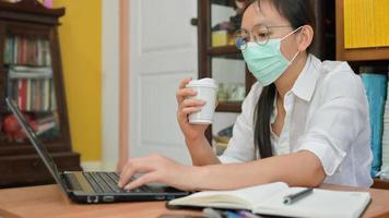 asiatische Frauenmaske, die eine Kaffeetasse hält und einen Laptop verwendet. Sie arbeitet zu Hause, um sich vor dem Koronavirus oder Covid-19 zu schützen.