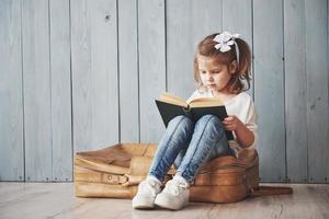bereit für große Reisen. glückliches kleines Mädchen, das ein interessantes Buch liest, das eine große Aktentasche trägt und lächelt. Reise-, Freiheits- und Vorstellungskonzept foto
