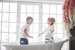 Kind spielt mit Rosenblättern im Badezimmer zu Hause. kleines mädchen und junge fawing spaß und freude zusammen. das Konzept der Kindheit und die Verwirklichung von Träumen, Fantasie, Vorstellungskraft