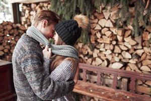 Winter-, Ferien-, Paar-, Weihnachts- und Menschenkonzept - lächelnder Mann und Frau in Hüten und Schal, die sich über hölzernem Landhaus und Schneehintergrund umarmen