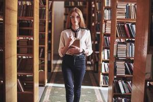junge attraktive Studentenbibliothekarin, die ein Buch zwischen den Bücherregalen der Bibliothek liest