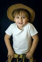 ein prägnant Porträt von ein europäisch Junge. Porträt auf ein dunkel Hintergrund von ein Junge im ein Cowboy Hut und ein Weiß T-Shirt. foto