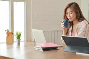 asiatisches studentenmädchen, das eine kaffeetasse hält und einen laptop verwendet, arbeitet an einem projekt, um ihr studium zu beenden. foto