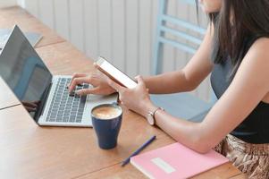 Frau, die ein Smartphone hält und einen Laptop auf einem Tisch mit einer Kaffeetasse an einem modernen Arbeitsplatz verwendet.