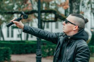 Blogger im Sonnenbrille Herstellung Selfie oder Streaming Video beim das Herbst Park mit Aktion Kamera mit kardanisch Kamera Stabilisator. gut aussehend Mann im schwarz Kleider Herstellung Foto