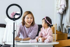 jung asiatisch Influencer Mutter ist Verkauf das Kinder Kleider Leben online Streaming beim Zuhause während erziehen ihr Kleinkind wie Hilfe foto
