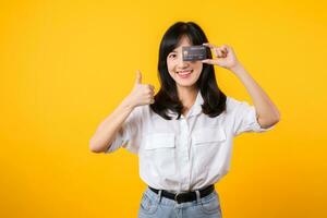 asiatisch jung Frau halten Anerkennung Karte im Vorderseite von einer Auge und zeigen Daumen oben Hand Geste mit glücklich Lächeln isoliert auf Gelb Hintergrund. Zahlung Einkaufen online Konzept. foto