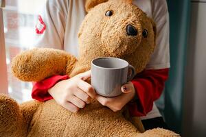 Nahansicht von Generation z Teenager Mädchen Trinken heiß Getränk durch Fenster mit Teddy Bär foto