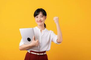 Porträt schön jung asiatisch Frau Unternehmer glücklich Lächeln tragen Weiß Hemd und rot Pflanzen zeigen zuversichtlich Körper Sprache Geste und halten Laptop Isolierung auf Gelb Hintergrund. foto