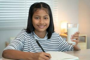 Kinderfrauen trinken Milch, um den Körper zu ernähren und das Gehirn zu ernähren. asiatische junge kleine Mädchen lernen zu Hause. mädchen trinken gerne milch und lesen ein buch für die prüfung, homeschool. Bildung, Vitamine, Entwicklung foto