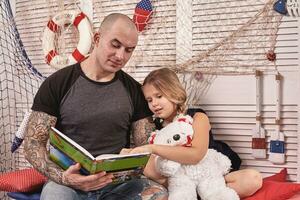 gut aussehend tätowiert Mann ist Ausgaben Zeit mit seine wenig süß Tochter. lesen Märchen während Tochter ist Sitzung nahe. glücklich Familie. foto