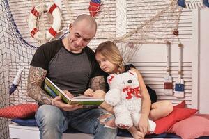 gut aussehend tätowiert Mann ist Ausgaben Zeit mit seine wenig süß Tochter. lesen Märchen während Tochter ist Sitzung nahe. glücklich Familie. foto