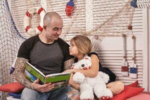 gut aussehend tätowiert Mann ist Ausgaben Zeit mit seine wenig süß Tochter. lesen Märchen während Tochter ist Sitzung nahe. glücklich Familie. foto