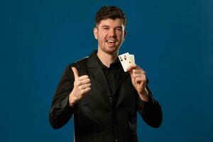 Neuling im Poker, im schwarz Weste und Shirt. halten zwei spielen Karten während posieren gegen Blau Studio Hintergrund. Glücksspiel, Kasino. Nahansicht. foto