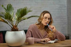 Mädchen Blogger im Rosa Strickjacke und Brille. Sitzung beim hölzern Tisch, gehen zu schreiben etwas im Notizbuch. Palme Baum im Topf, Tasse und Tablette in der Nähe foto